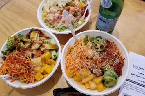 Three colorful bowls of fresh salads with various toppings, alongside a bottle of sparkling water and a menu.
