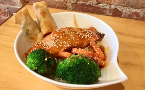 A bowl of sesame-glazed salmon with broccoli, carrots, and spring rolls on a wooden table.