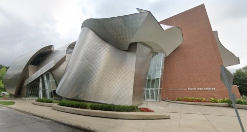 Modern architectural building with a unique, curved metallic facade and brick elements, surrounded by landscaped grounds.