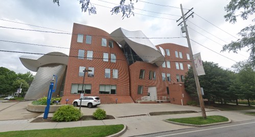A modern, wavy architectural building with large windows, surrounded by greenery and a parking area.