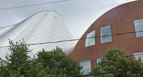 Modern building with a curved metallic roof and a brick facade, surrounded by trees and power lines.