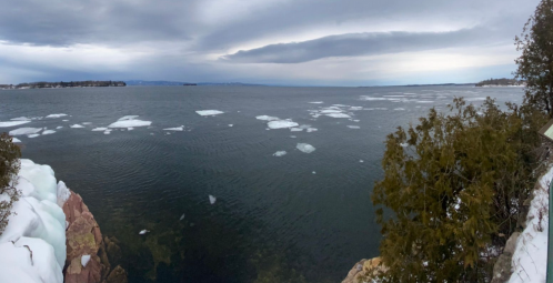 Panoramic view of a calm lake with ice patches, surrounded by rocky shores and trees under a cloudy sky.