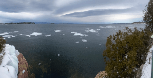 Panoramic view of a calm lake with ice patches, surrounded by rocky shores and trees under a cloudy sky.