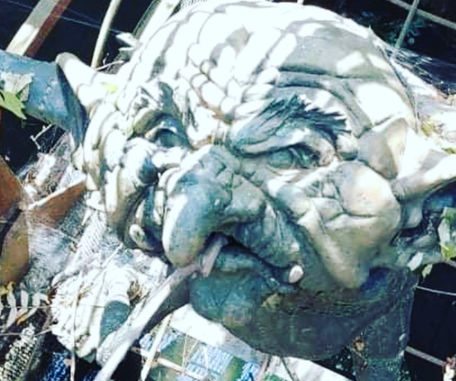 A close-up of a whimsical, weathered goblin sculpture surrounded by leaves and debris.