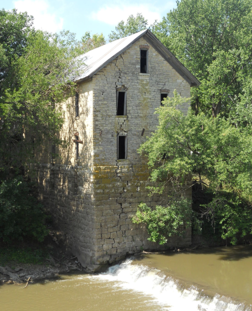 Abandoned stone building beside a river, partially hidden by trees, with a small waterfall in the foreground.