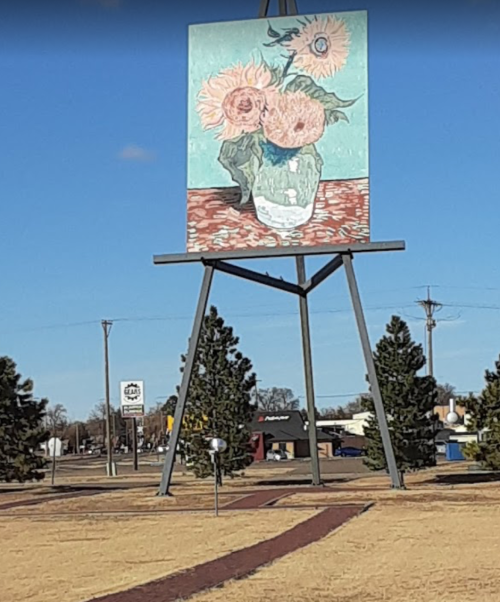 A large sign displays a painting of sunflowers in a vase, set against a clear blue sky and grassy landscape.