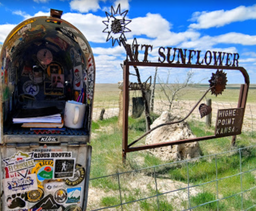 A decorated mailbox next to a sign for Mt. Sunflower, the highest point in Kansas, against a blue sky.