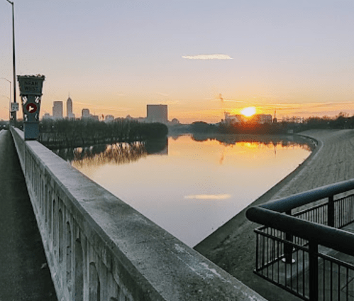 Sunset over a calm river with city skyline in the background, viewed from a walkway beside the water.