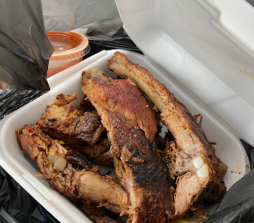 A takeout container with several pieces of grilled ribs and a small cup of sauce on the side.
