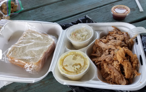 A takeout container with pulled pork, coleslaw, potato salad, and a slice of bread, accompanied by a small sauce cup.