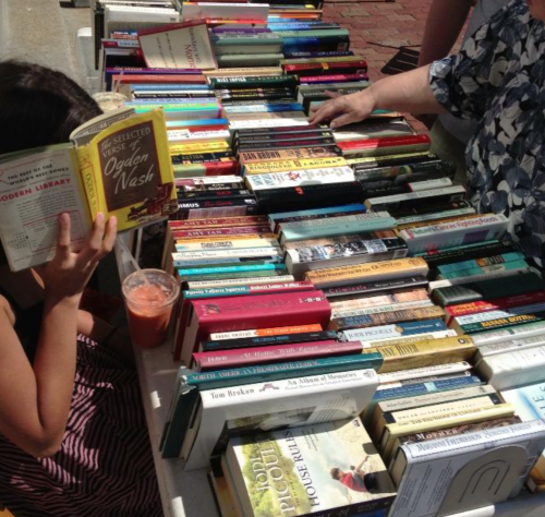 A person sits at a table with books and a drink, surrounded by stacks of books in an outdoor setting.