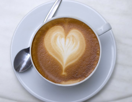 A cup of coffee with a heart-shaped latte art design on top, placed on a white saucer with a spoon.