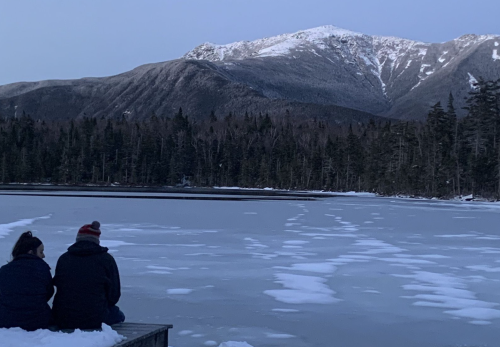 Two people sit on a dock by a frozen lake, surrounded by snow-covered mountains and trees.