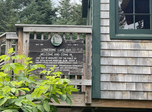 Sign for Lonesome Lake Hut, welcoming visitors, with wooden structure and greenery in the foreground.