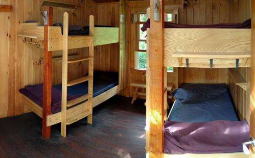 Cozy wooden cabin interior with two sets of bunk beds and a single bed, featuring warm wood paneling and natural light.