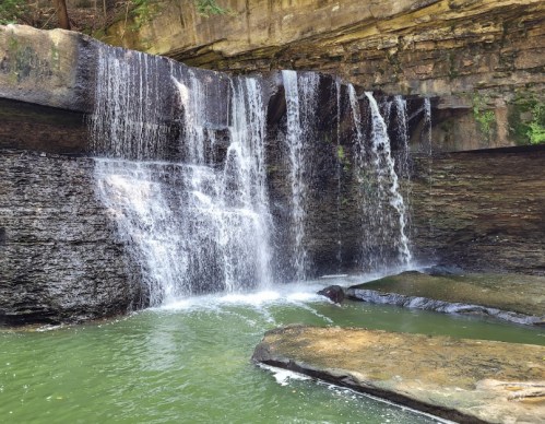 A serene waterfall cascading over rocky cliffs into a calm green pool below, surrounded by lush greenery.