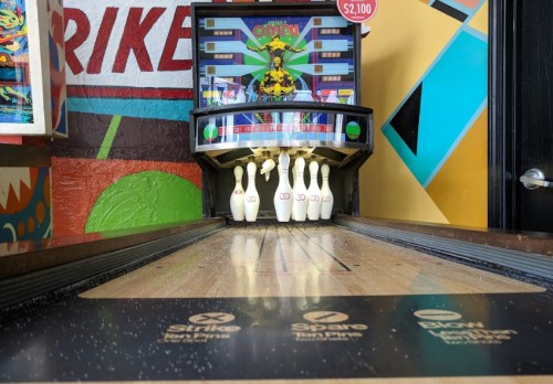 A bowling alley lane with pins set up, colorful wall art in the background, and a retro bowling game machine.