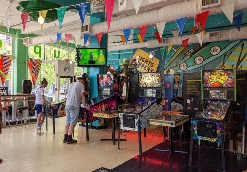 A vibrant arcade with colorful decor, featuring pinball machines and people playing games. Banners hang from the ceiling.