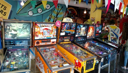 A colorful arcade with various pinball machines lined up, featuring vibrant artwork and decorations in the background.