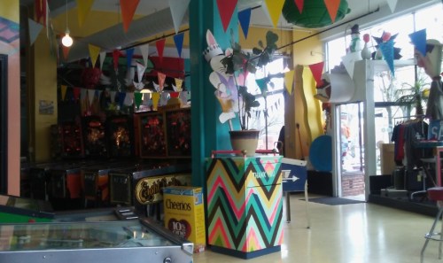 Colorful arcade interior with pinball machines, vibrant decorations, and a potted plant in the foreground.