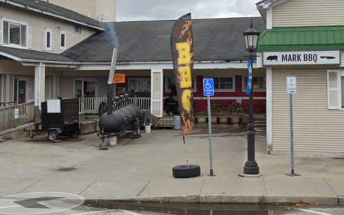 A BBQ restaurant with a large smoker outside, a "BBQ" flag, and a parking area.