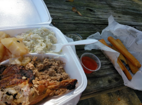 A takeout meal featuring grilled chicken, pulled pork, coleslaw, mashed potatoes, and breadsticks on a wooden table.