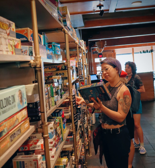 A person with purple hair examines a board game on a shelf in a cozy store, while another person browses nearby.