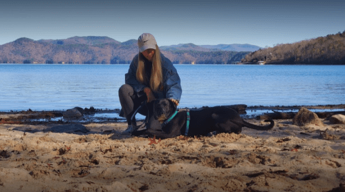 A person kneels on a sandy beach beside a black dog, with mountains and water in the background.