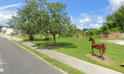 A grassy area with metal horse sculptures under a blue sky, surrounded by trees and a sidewalk.