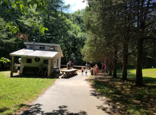 A path leads to a small building surrounded by trees, with people gathered outside enjoying a sunny day.