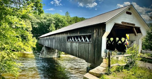 A historic covered bridge spans a river, surrounded by lush greenery and a bright blue sky.