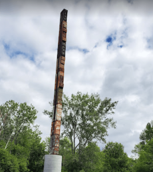 A tall totem pole stands among green trees under a cloudy sky.