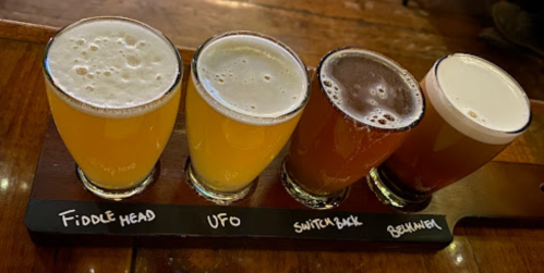 A wooden flight board with four glasses of different craft beers, labeled Fiddle Head, UFO, Switchback, and Brehm.