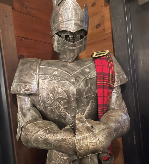 A metallic knight statue wearing armor and a red tartan sash, standing against a wooden wall.