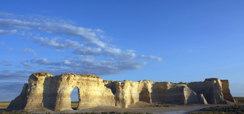 A vast landscape featuring a large rock formation with a natural arch, under a clear blue sky and scattered clouds.