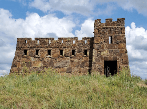 A stone castle ruin with a tower, set against a backdrop of blue sky and fluffy clouds. Grass surrounds the structure.