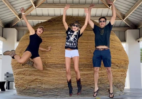 Three people jump joyfully in front of a large haystack, wearing casual summer outfits and smiling.