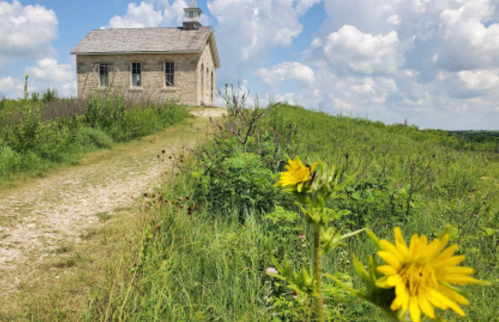 A stone building on a grassy path, with a bright yellow flower in the foreground and a blue sky with clouds above.