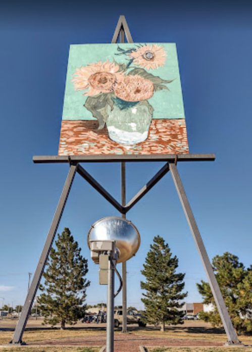 A large easel displays a painting of sunflowers against a turquoise background, with trees in the background.