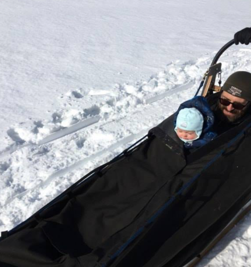 A person pulls a sled with a baby bundled up in winter gear across a snowy landscape.