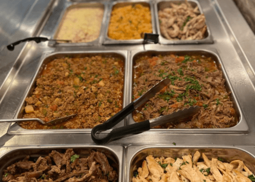 A buffet display with various dishes, including seasoned meats, rice, and vegetables in metal trays.