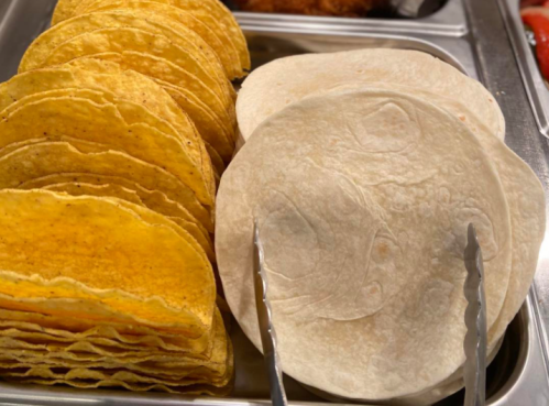 A tray of stacked yellow corn tortillas and white flour tortillas, with tongs resting on the side.