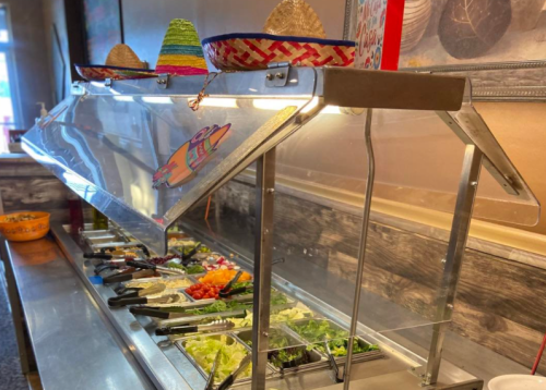 A salad bar with fresh toppings and sombreros on display, under a glass cover in a restaurant setting.