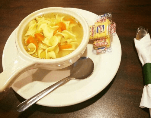 A bowl of chicken noodle soup with carrots, served with crackers and a spoon on a wooden table.