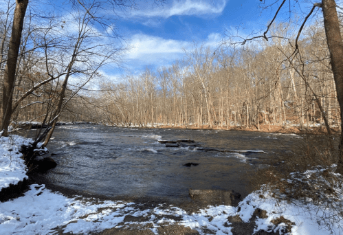 A serene river flows through a snowy landscape, surrounded by bare trees under a blue sky with scattered clouds.