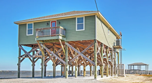 A green beach house on stilts, with a red door and balcony, overlooking a sandy area and water under a clear blue sky.