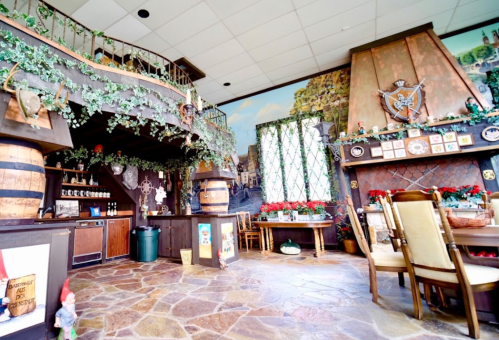 Cozy restaurant interior with wooden decor, greenery, and a stone floor, featuring a staircase and festive table settings.