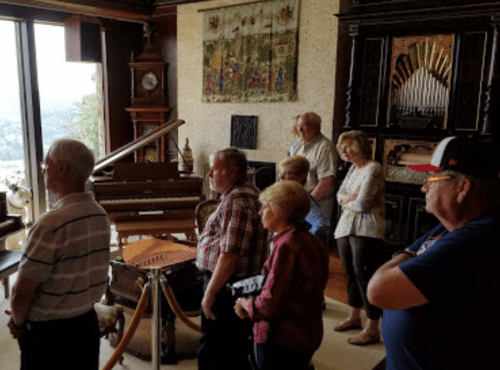 A group of people stands in a room with a piano and ornate decor, listening attentively to a presentation.