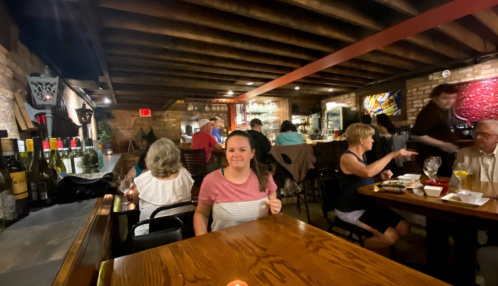 A woman sits at a table in a cozy restaurant, with wine bottles in the background and diners enjoying their meals.