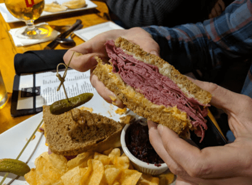 A person holds a thick pastrami sandwich with rye bread, accompanied by pickles and chips on a plate.
